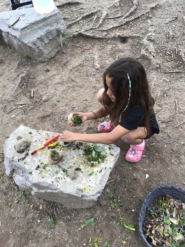 One child crouching beside a large rock with crushed green leaves and grass, the child is pinching some of the green