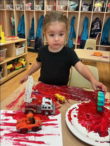 Toddler standing at the table with paint on paper, cars on paper with tracks of paint and a plate full of paint
