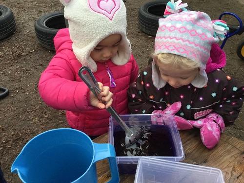 Two preschool girls are stirring water with food colouring in it while outside.