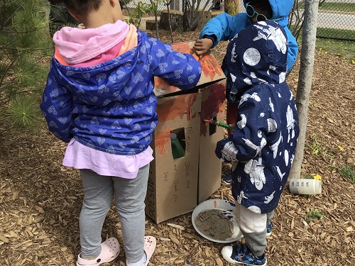 Group of children painting the cardboard house together.
