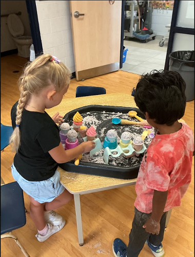 2 summer camp children standing at the tuff tray with sand, scoops, ice cream cones and trays facing away from the camera and towards the tuff tray