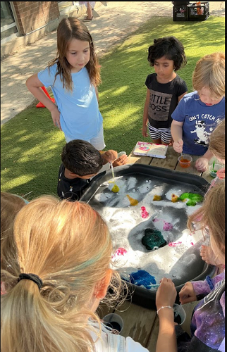 8 summer camp children standing around and looking at the tuff tray filled with bubbling water, one summer camp child is holding a pipette and holding it over some yellow