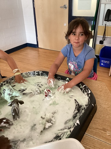 summer camp child sitting at the table with a tuff tray full of oobleck and animals, the child has her hands in the oobleck on the tuff tray